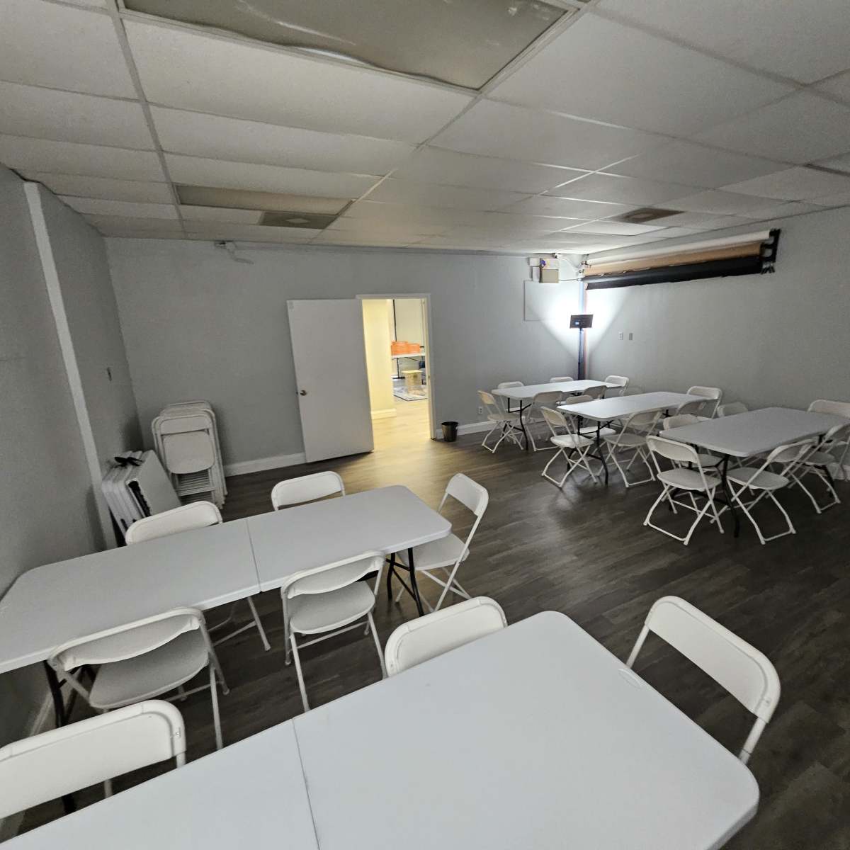 A large, empty room arranged with multiple rectangular tables and folding chairs, featuring a doorway leading to another space.