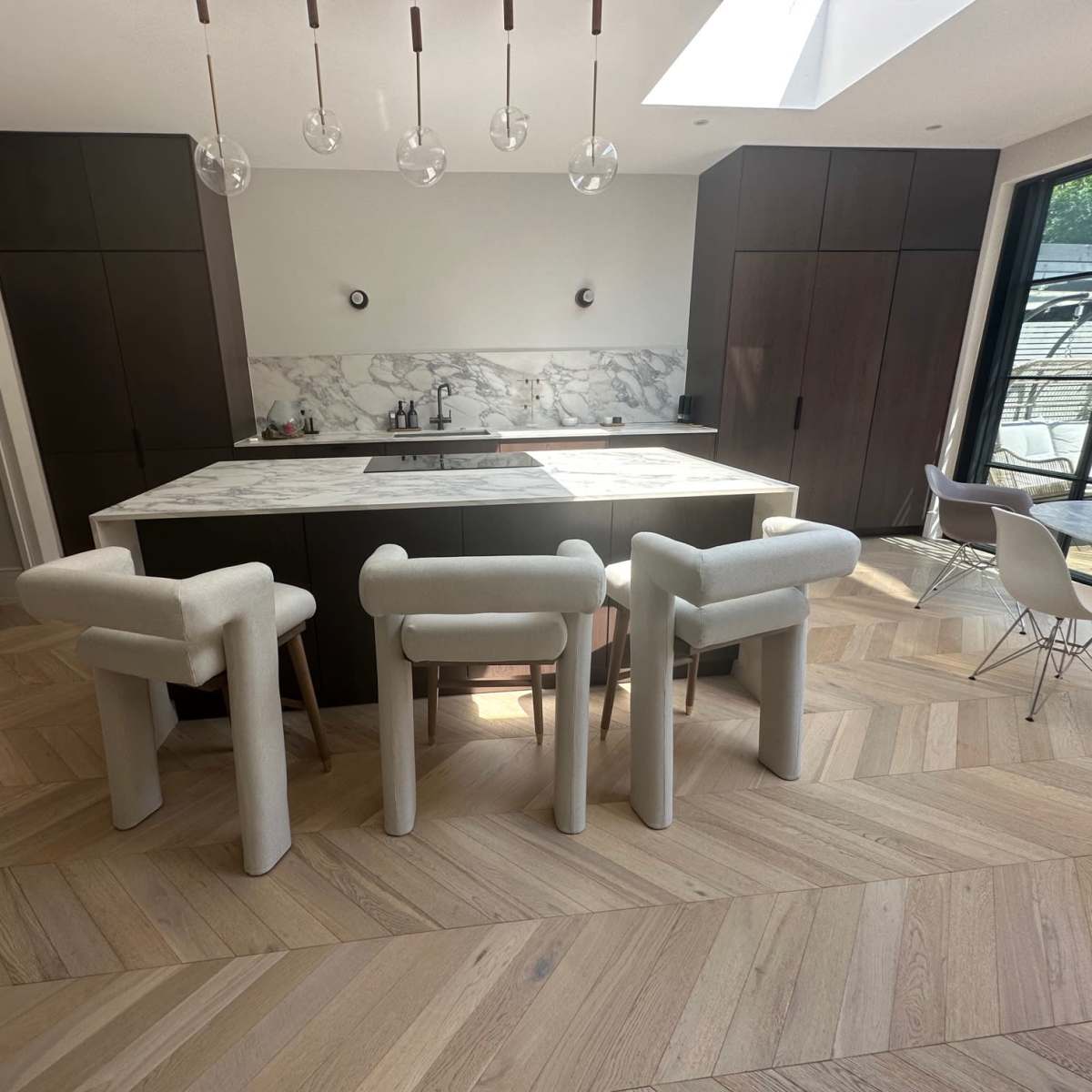A modern kitchen with a marble countertop, dark cabinetry, and three upholstered chairs arranged around an island, all set on herringbone-patterned wooden flooring.