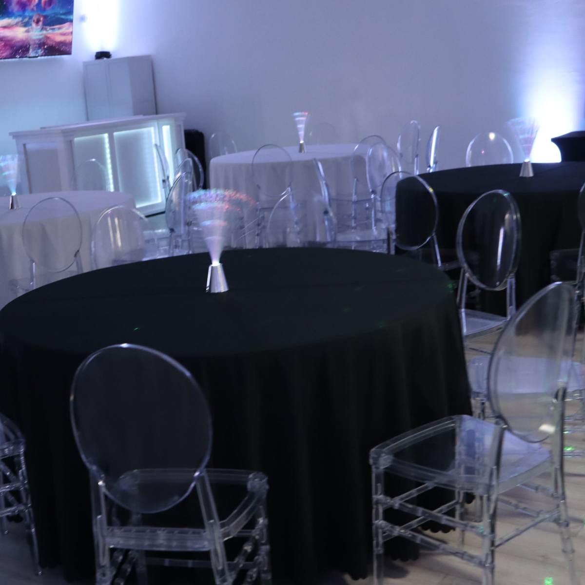 An empty event space with round tables covered in black and white tablecloths, surrounded by transparent chairs.