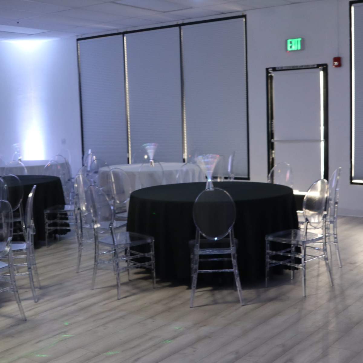 A spacious event room with several round tables covered in black and white linens, surrounded by clear acrylic chairs, and illuminated by soft lighting.