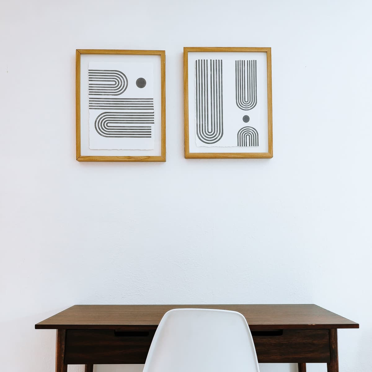 A wooden desk with a white chair is positioned against a white wall, featuring two framed geometric prints above it.
