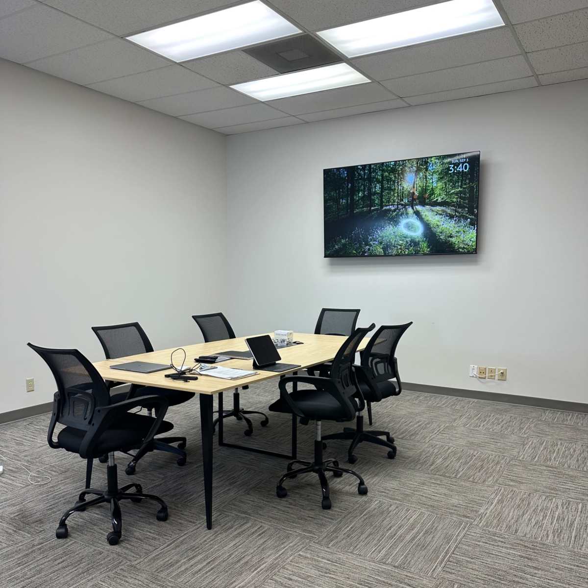 A modern conference room features a rectangular table with six black chairs, a laptop, and a television displaying a forest scene on the wall.