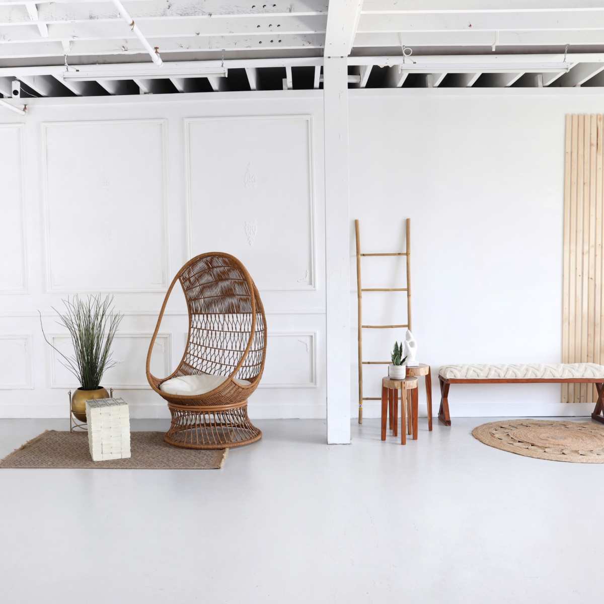 A minimalist interior space featuring a rattan chair, a mirror, various decorative plants, and several wooden furniture pieces against a white wall.