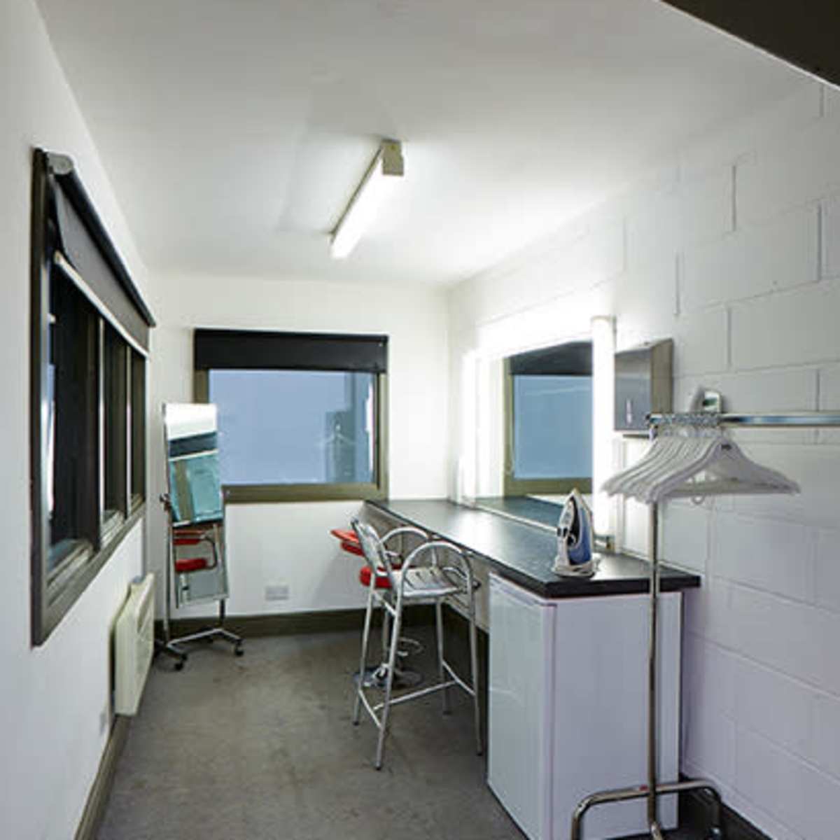 A small, minimalist dressing room with a countertop, two mirrors, a clothing rack, and a few chairs.