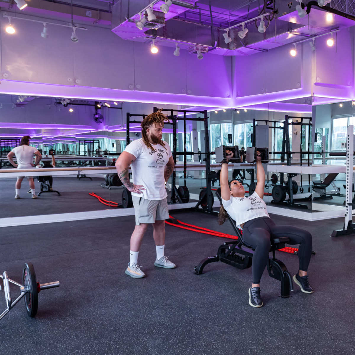 A person lifts weights on a bench while a trainer observes in a gym with purple lighting and workout equipment.