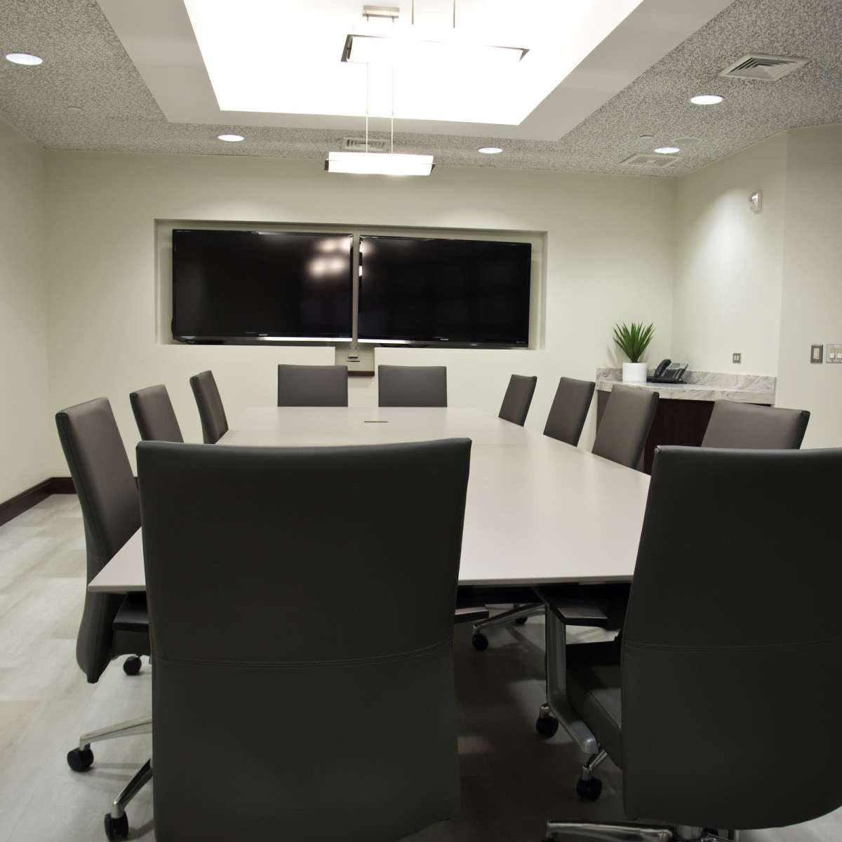 A modern conference room features a rectangular table surrounded by ten gray chairs, with two large screens mounted on the wall and framed art on the adjacent walls.