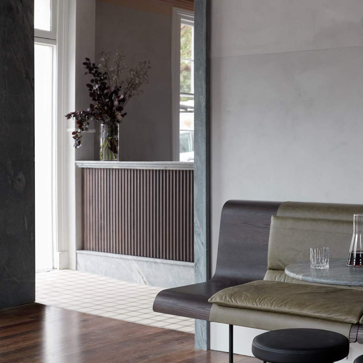 A minimalistic interior features a grey wall, a dark wooden bench with a round table, and a reception area with a marble counter in the background.