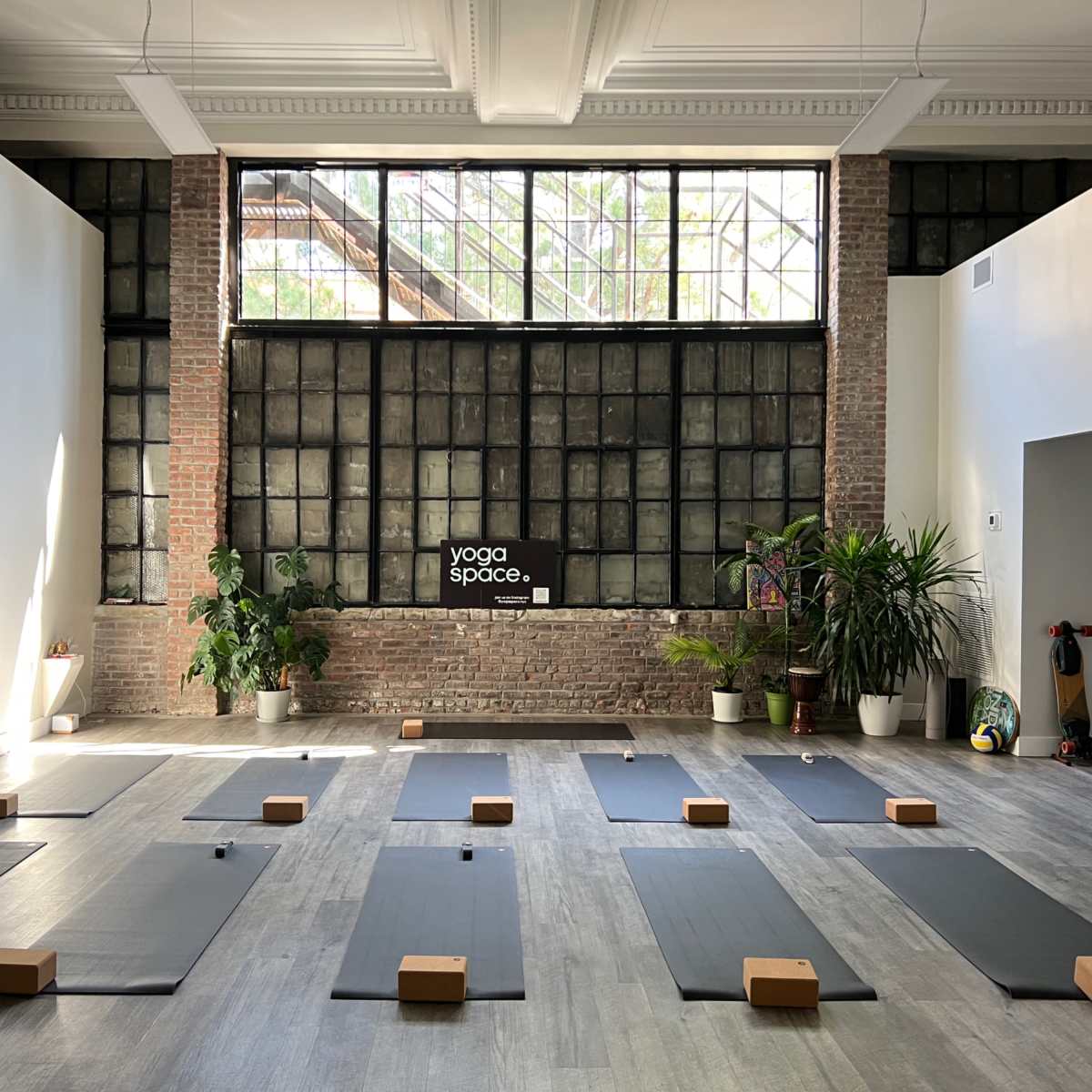 A yoga studio with several yoga mats arranged in rows, wooden blocks placed on each mat, and large windows letting in natural light.