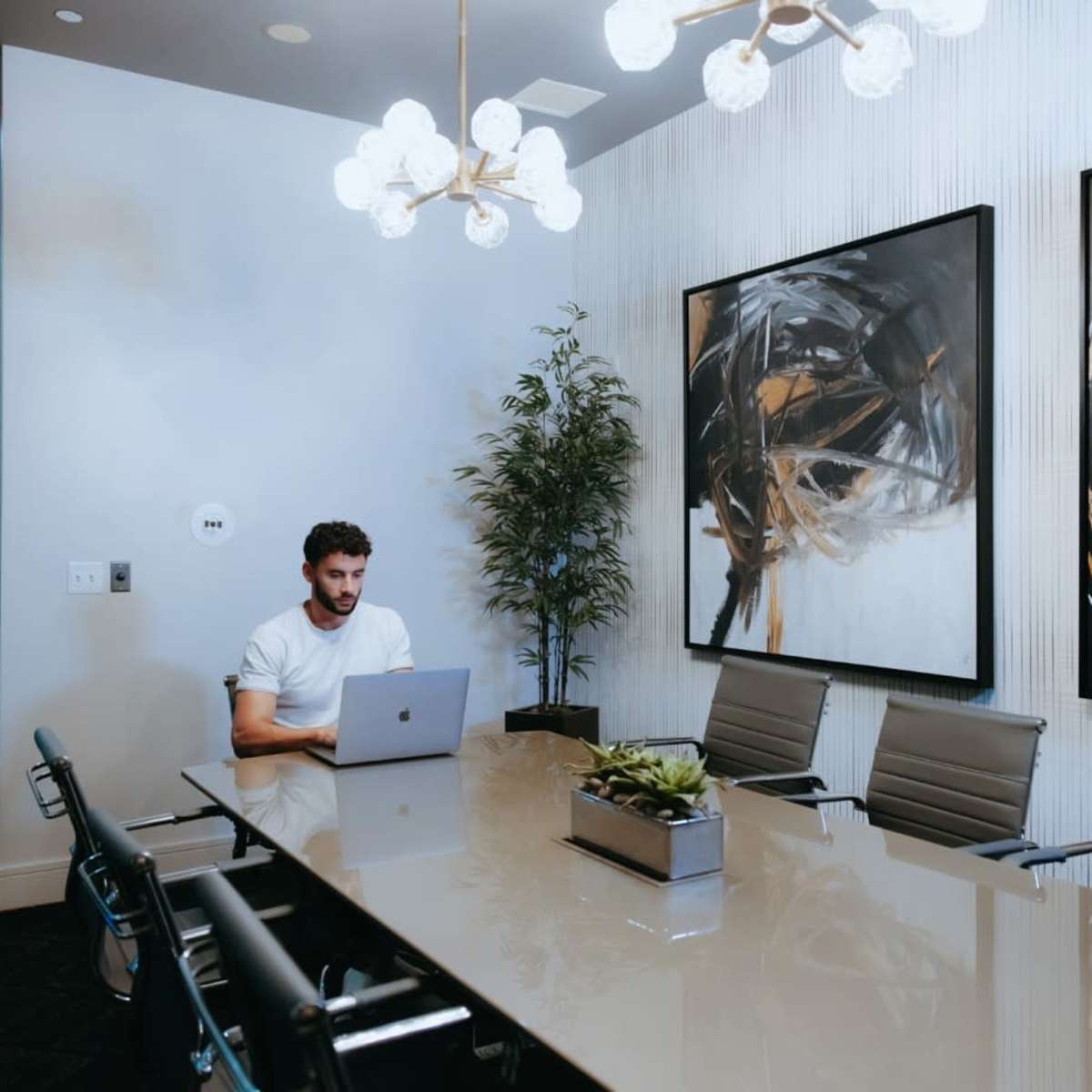 A man sits at a conference table with a laptop in a well-lit meeting room featuring abstract artwork on the walls.