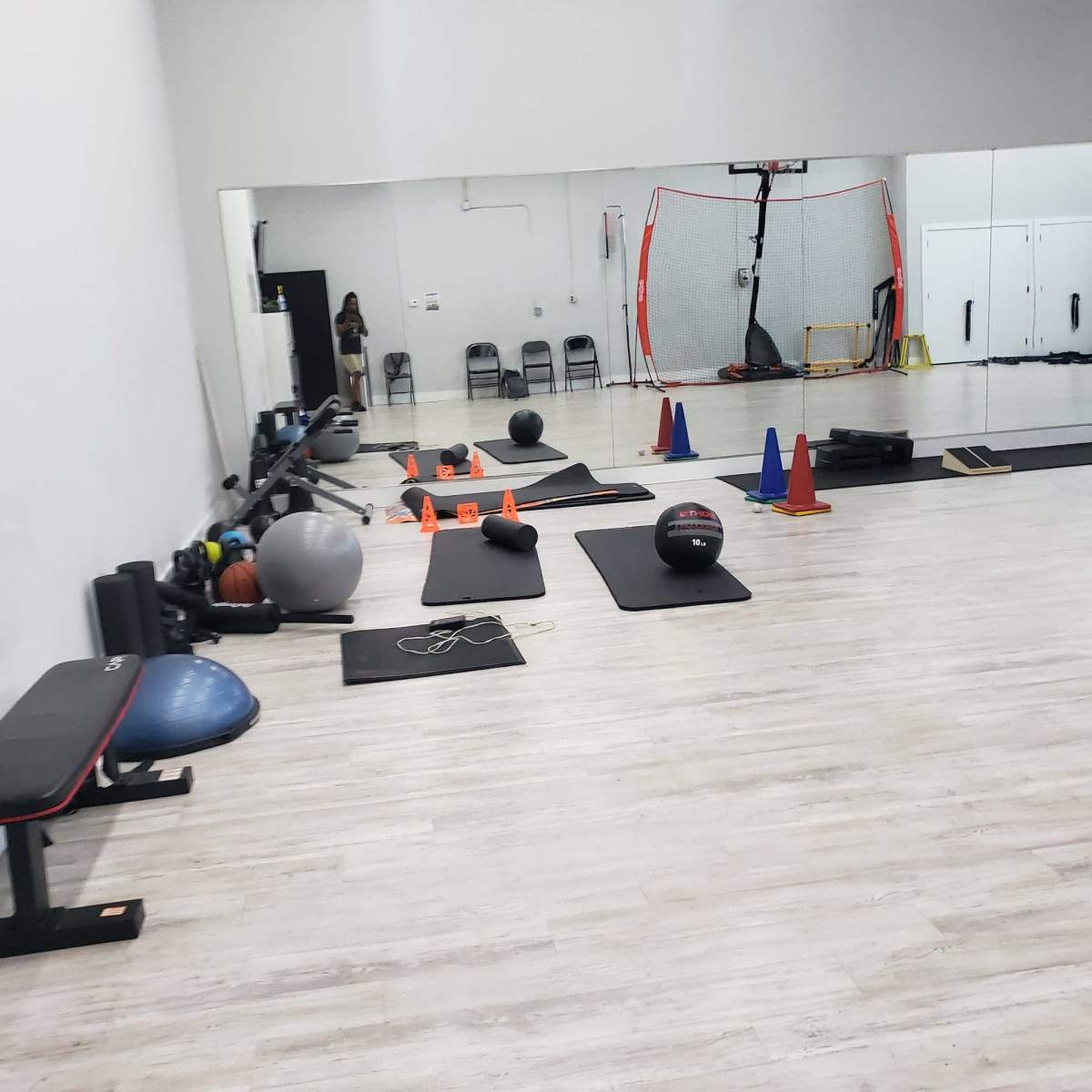 A gym interior with exercise equipment, including mats, exercise balls, cones, and a basketball hoop visible in the background.