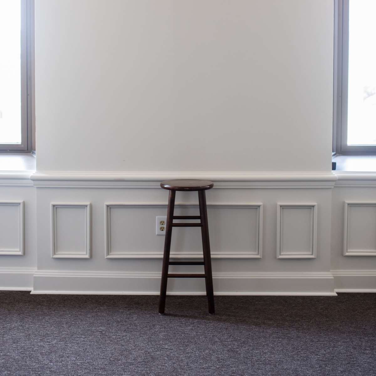 A wooden stool stands against a blank wall between two windows in a room with wainscoting and carpeted flooring.