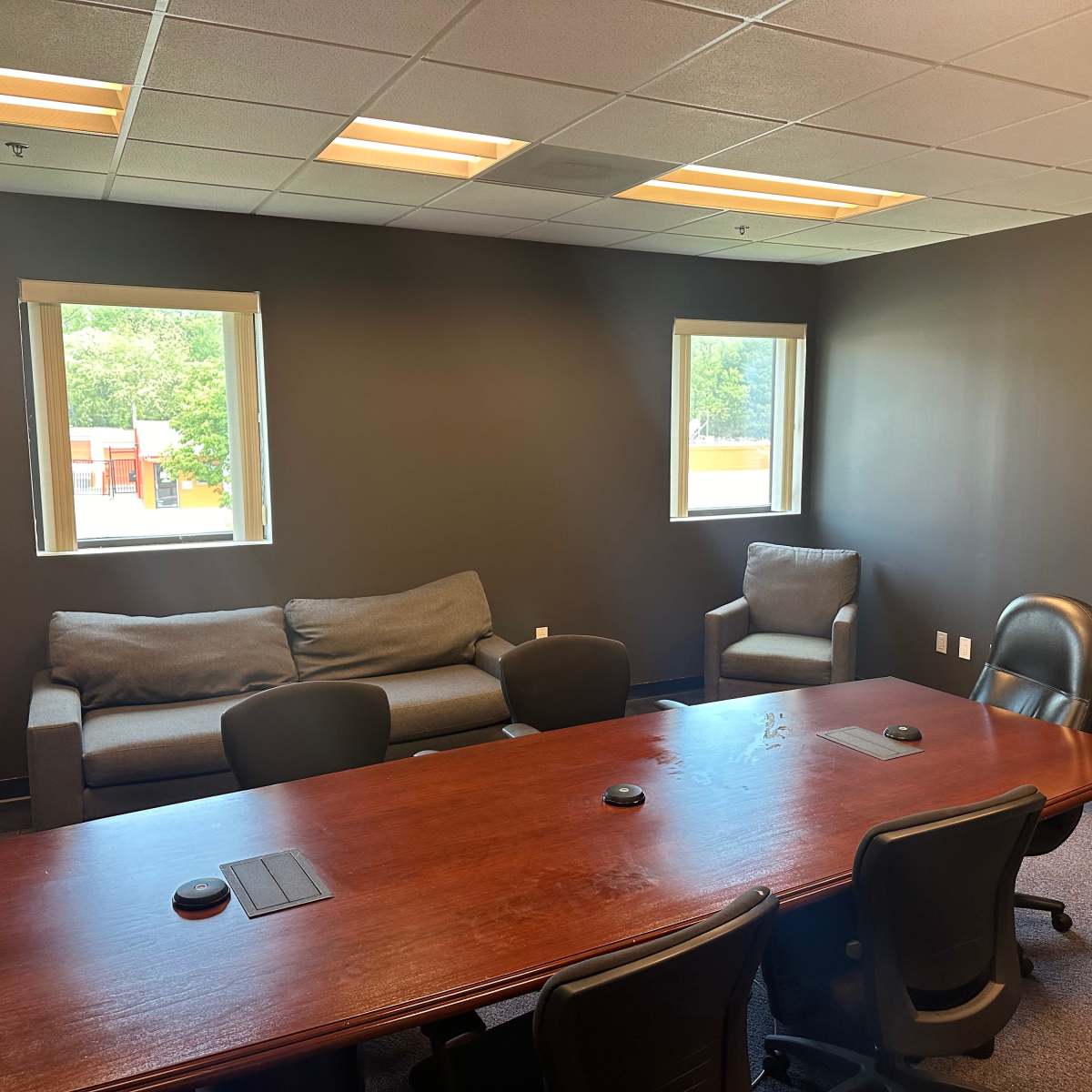 A conference room featuring a wooden table surrounded by black chairs, with a couch and an armchair against the wall and two windows letting in natural light.