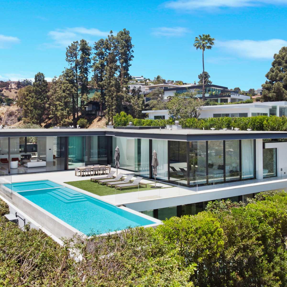 A modern house with extensive glass windows features a swimming pool and is surrounded by greenery and hills.