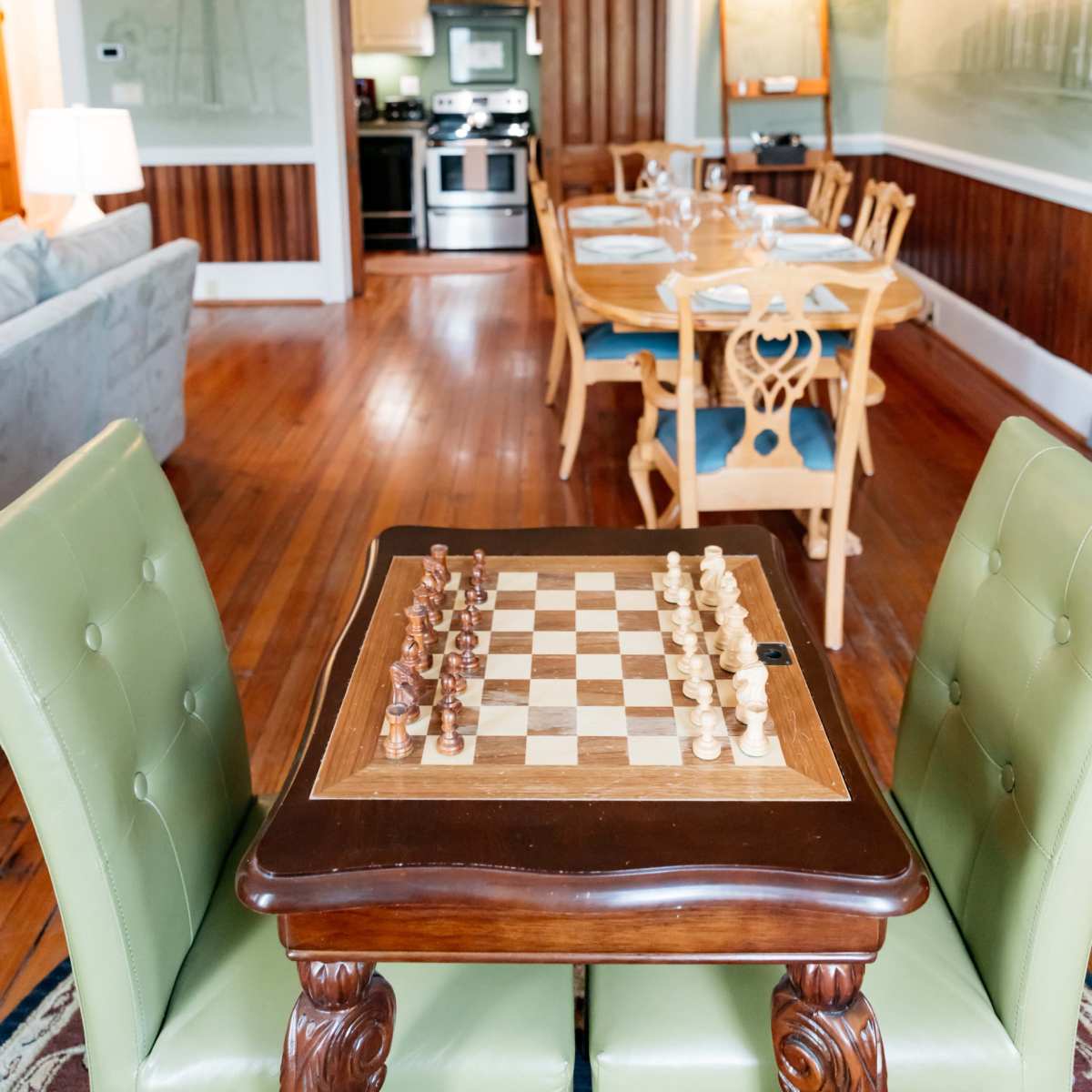 A chessboard sits on a wooden table in the foreground, with a dining area and kitchen visible in the background.