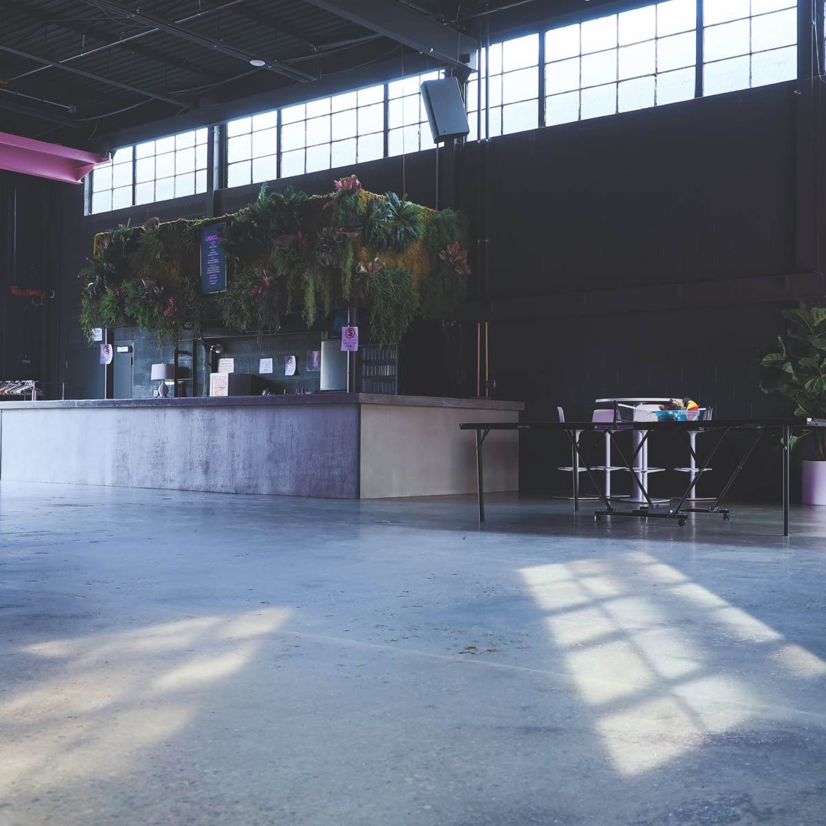 A spacious interior with a bar area featuring a lush vertical garden, surrounded by tables and chairs under large windows allowing natural light to enter.