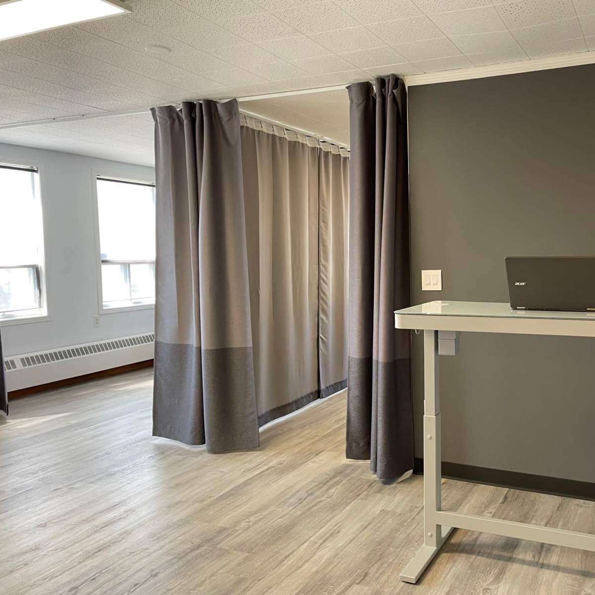 A modern workspace featuring a standing desk with a laptop and a small potted plant, adjacent to a curtained area in a room with light wood flooring and large windows.