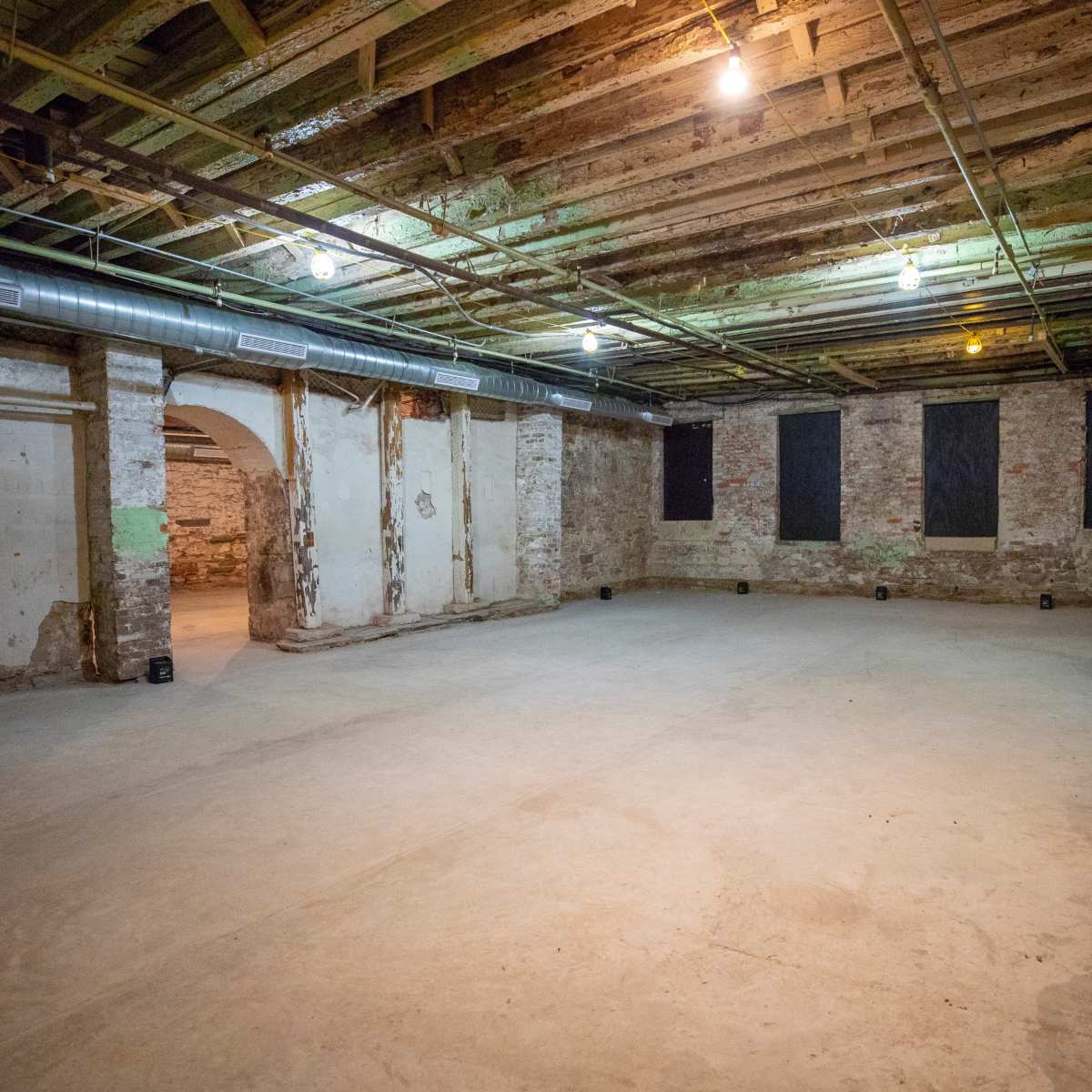 A spacious, empty basement with exposed wooden beams and stone walls, illuminated by overhead lights.