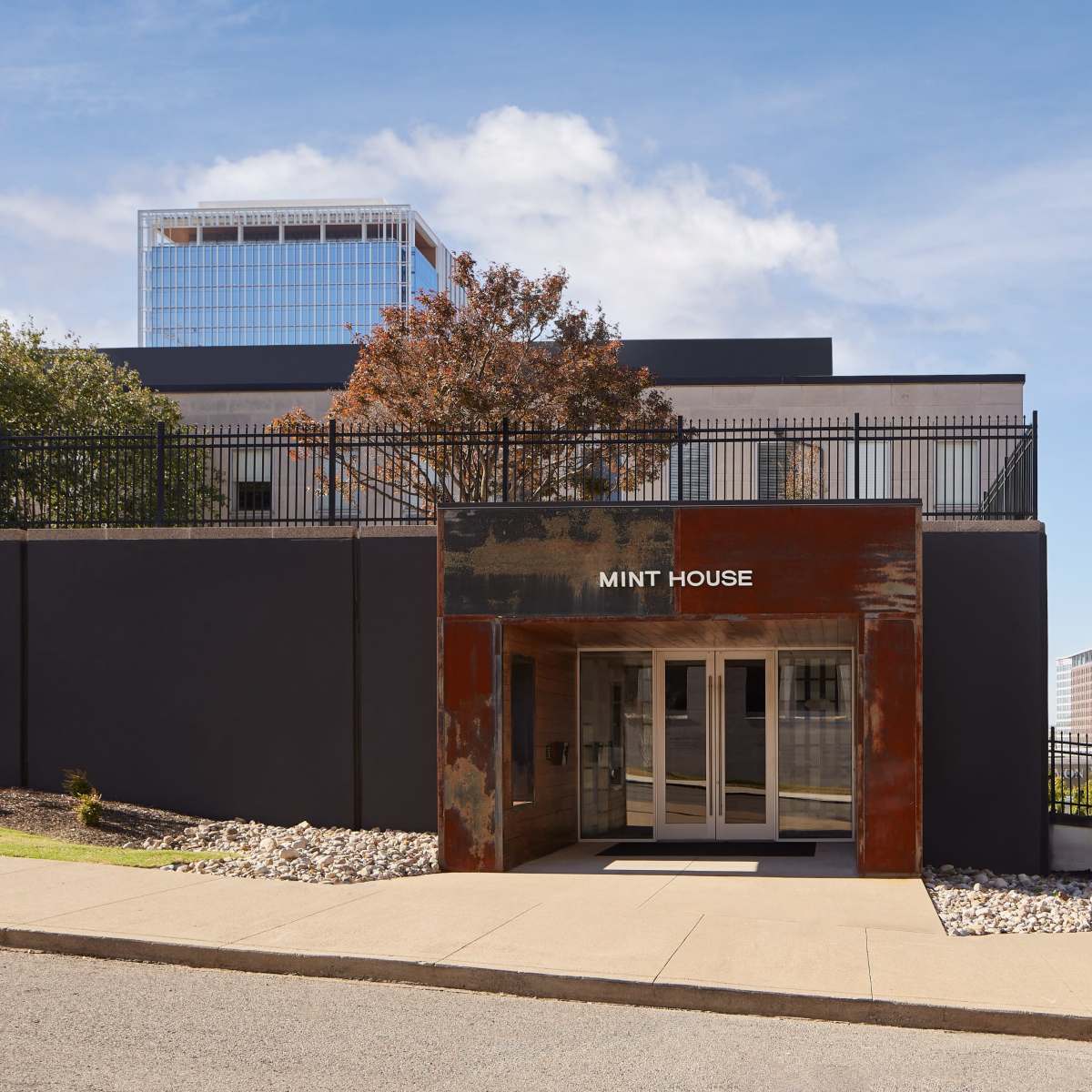 The entrance of a modern building named "Mint House" features a rusted metal facade and large glass doors, set against a clear blue sky.