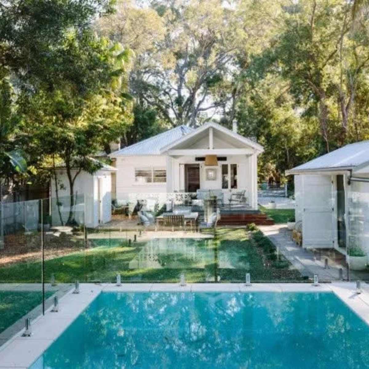 A clear swimming pool is situated in front of a modern house surrounded by greenery and a glass fence.