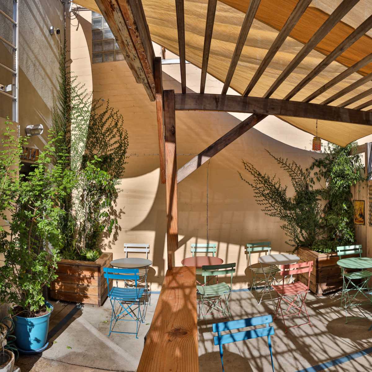 A patio area with colorful metal tables and chairs surrounded by greenery and shaded by a striped canopy.