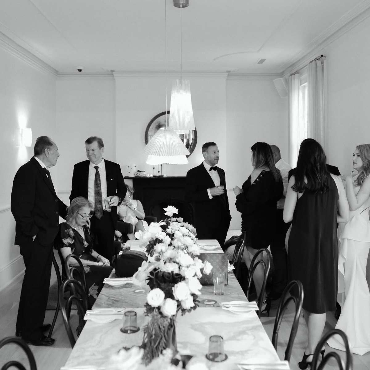 A group of elegantly dressed individuals socializes around a dining table adorned with flowers in a well-lit room.