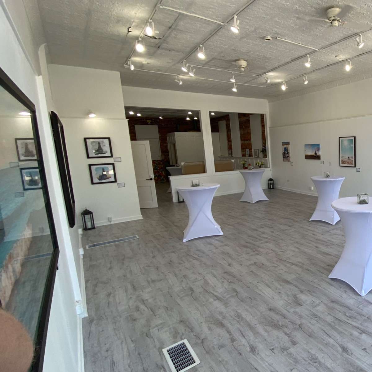 A spacious, well-lit gallery featuring several white cocktail tables, framed photographs on the walls, and a chalkboard sign promoting a grand opening event.
