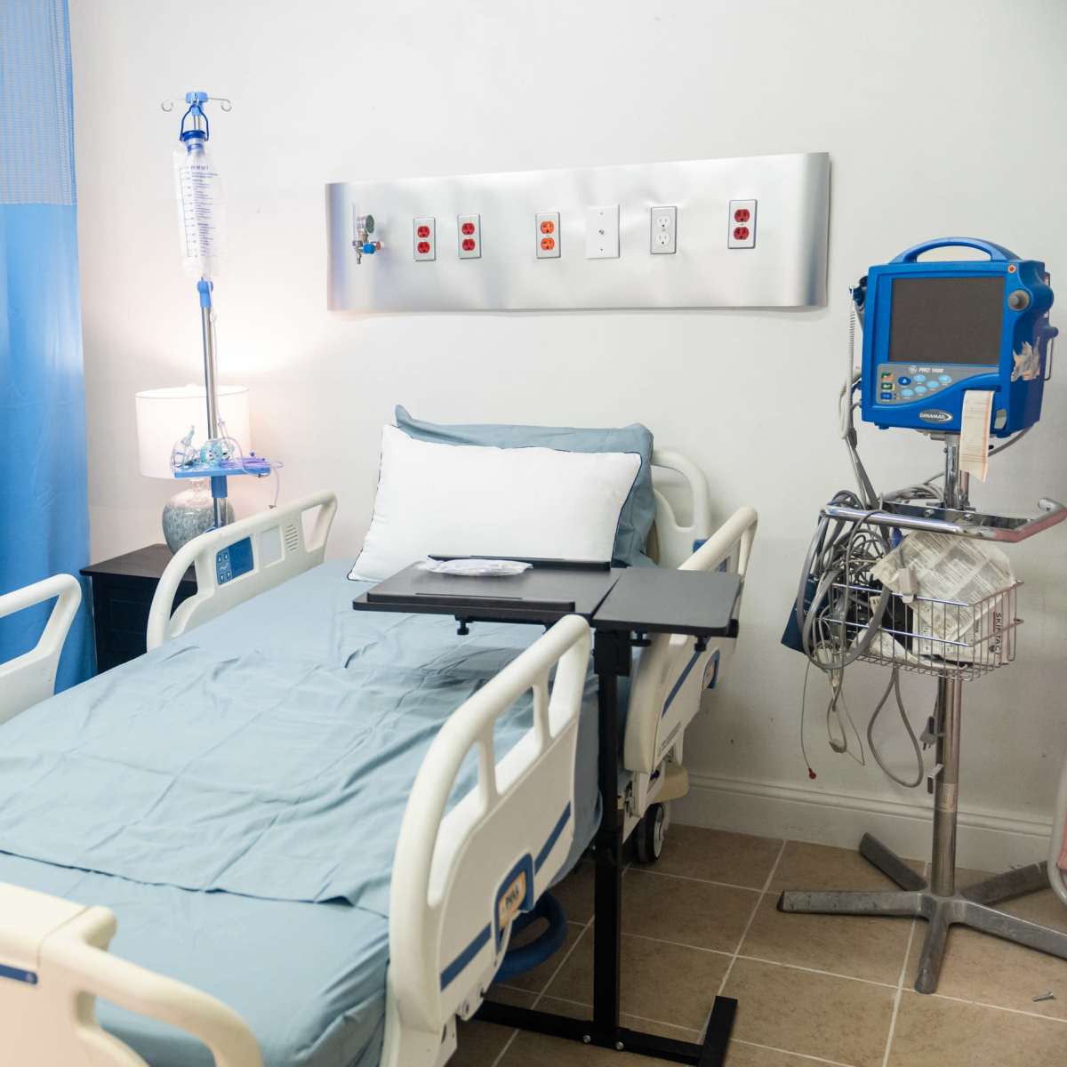 A hospital room featuring a bed with light blue linens, a bedside table, and various medical equipment along the wall.