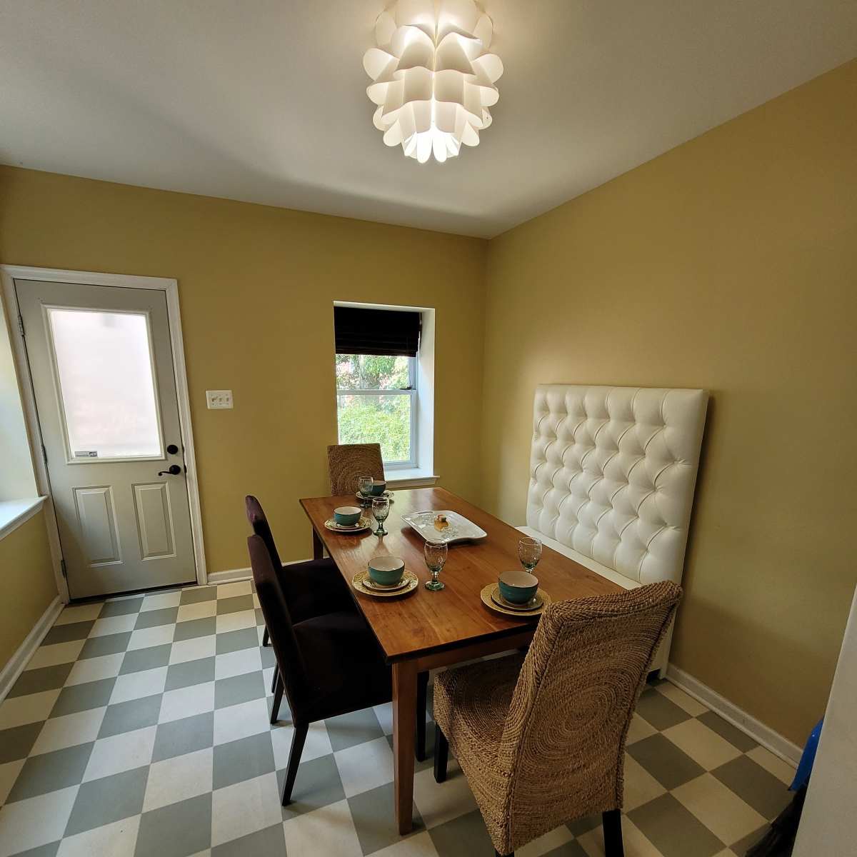 A dining area features a wooden table set for four, with a white tufted wall panel, minimalist light fixture, and checkered floor.