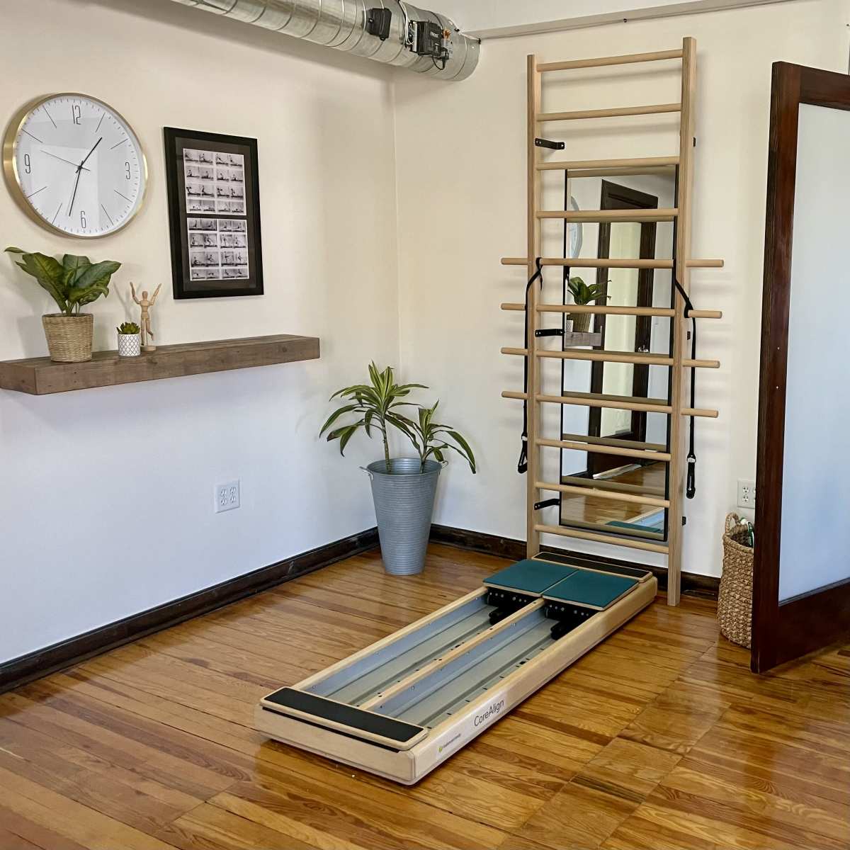 A fitness studio with a reformer machine on wooden flooring, a wall-mounted ladder, a mirror, a clock, and decorative plants.