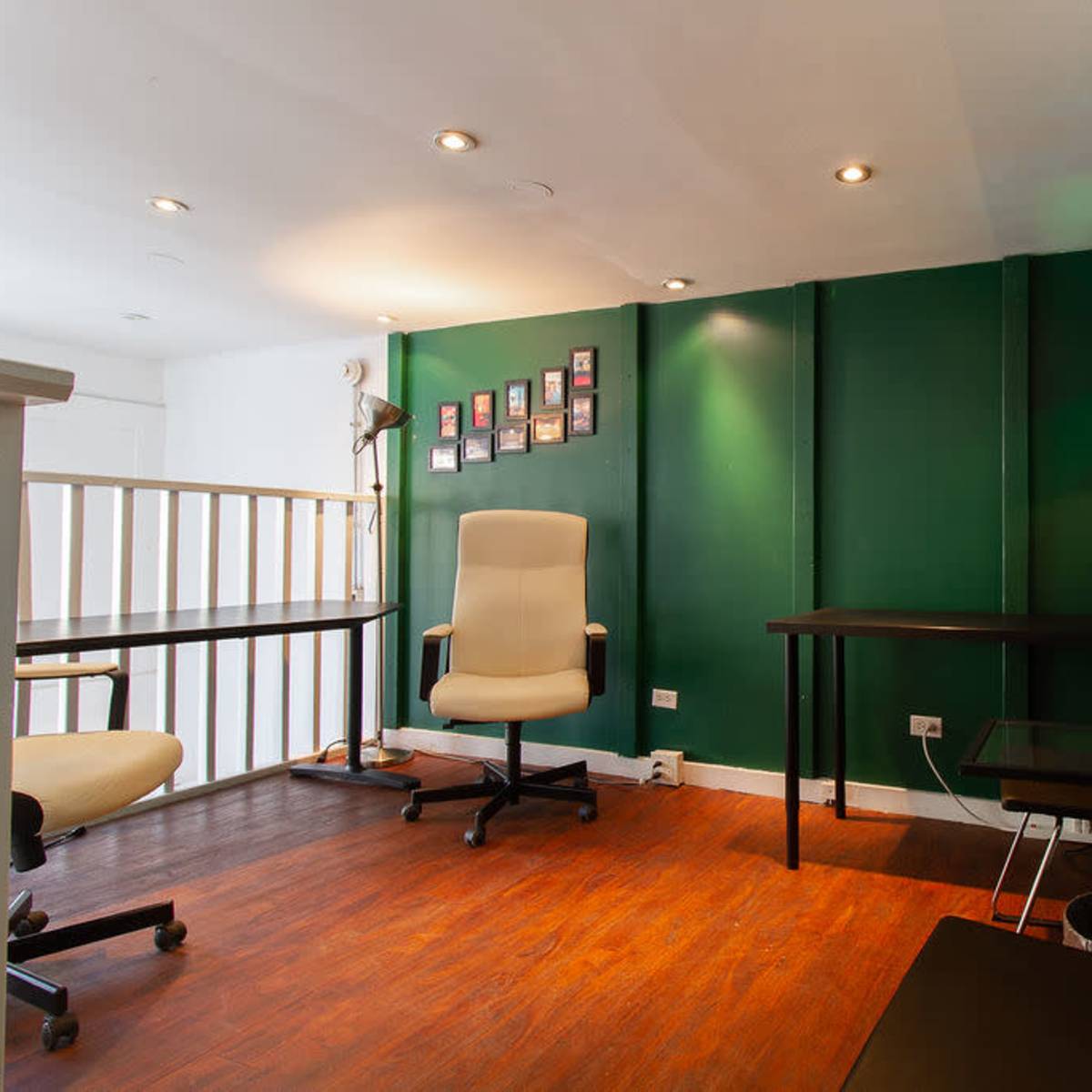 A small office space with green walls, wooden flooring, a desk, an office chair, and a wall displaying several framed photographs.