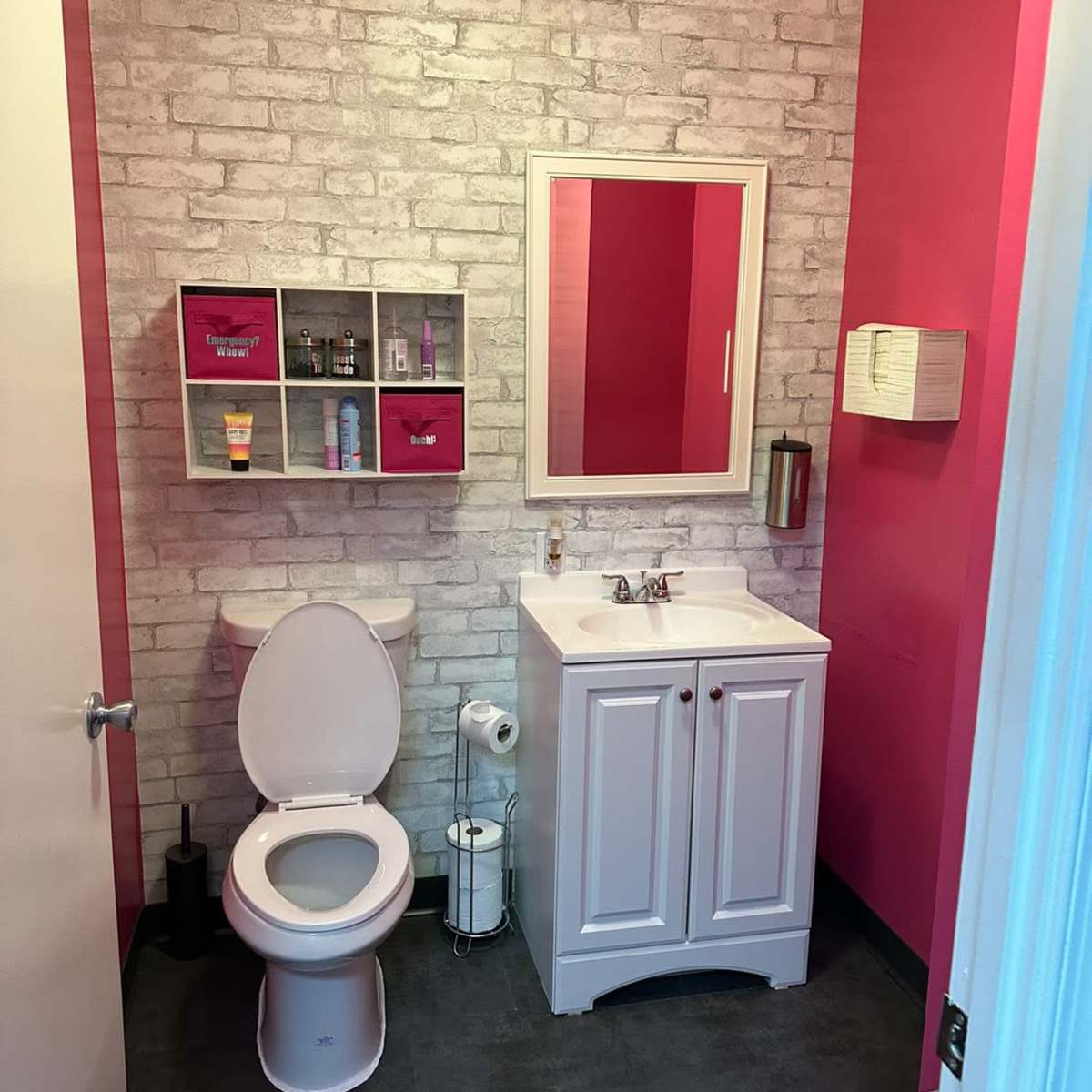 A bathroom featuring a white toilet, a white sink cabinet, a wall mirror, and pink accent walls with a brick design.