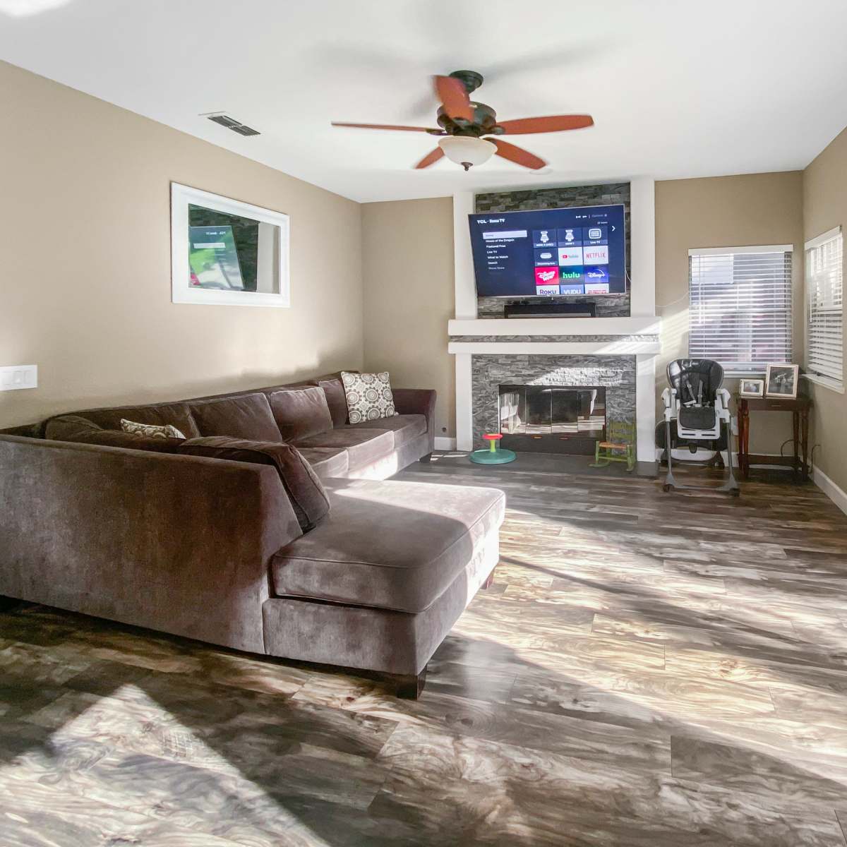 A living room with a dark sectional sofa, a stone fireplace, a large TV mounted on the wall, and sliding glass doors that lead outside, all set on a wooden floor.