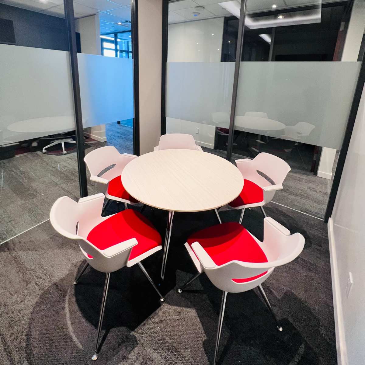A small conference room features a round table surrounded by six chairs with red cushions, enclosed by glass walls.