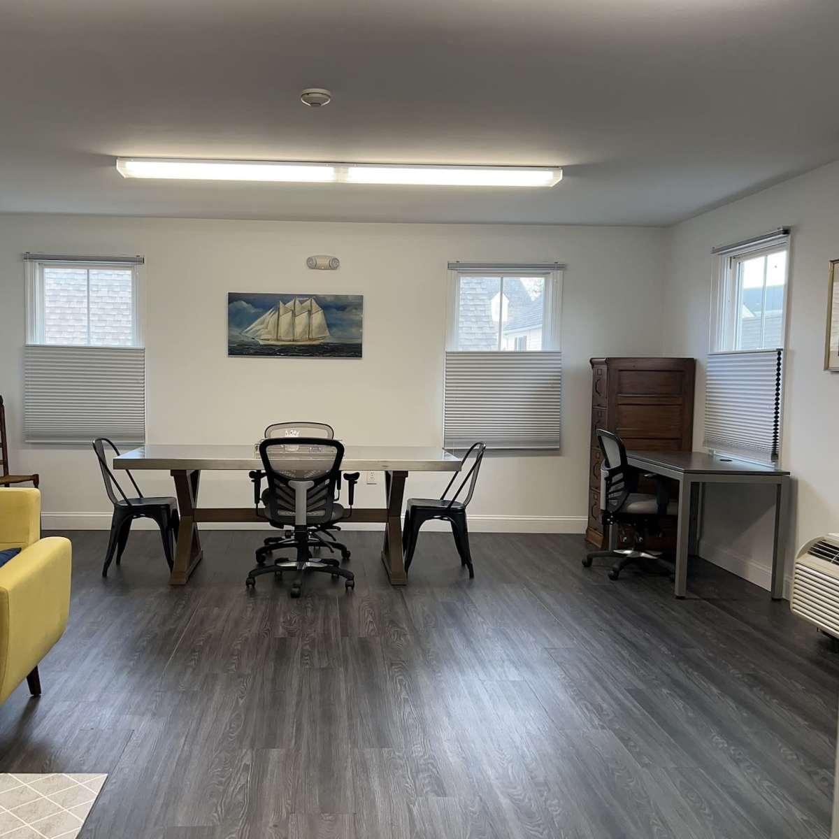 A bright room with a yellow couch, two desks, several chairs, and windows with blinds.