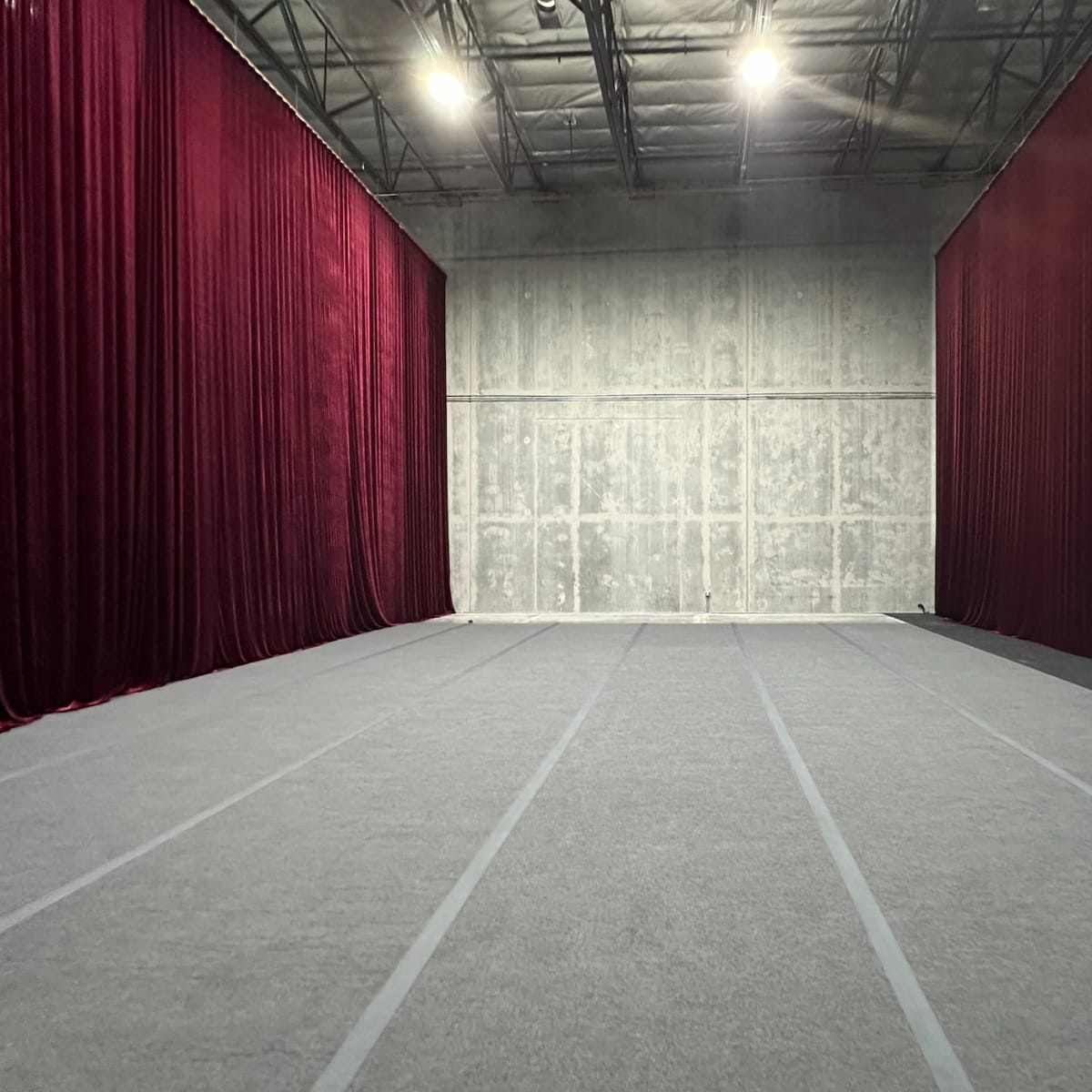 An empty space divided by deep red curtains, with a gray carpet and concrete walls.