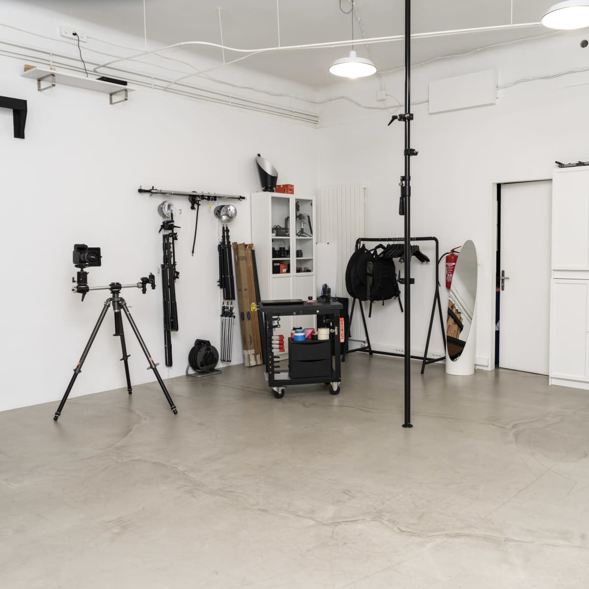 A minimalist studio space features a tripod-mounted camera, a portable cart with equipment, and various backdrops against white walls.