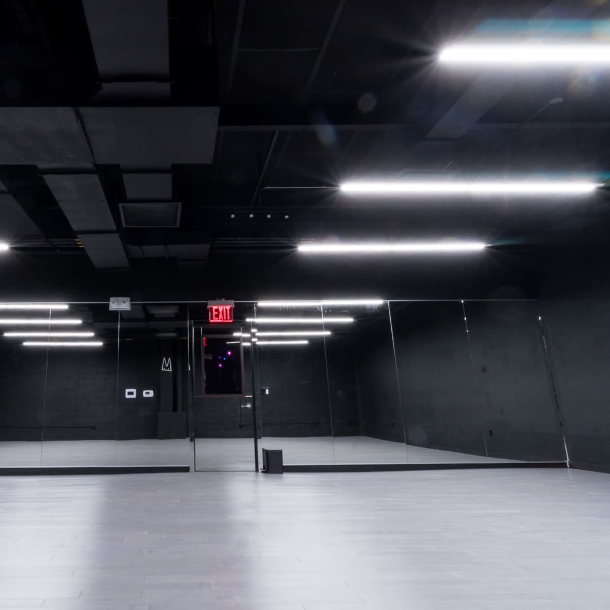 A sparsely furnished black dance studio with bright overhead lights and large mirrors on one wall.