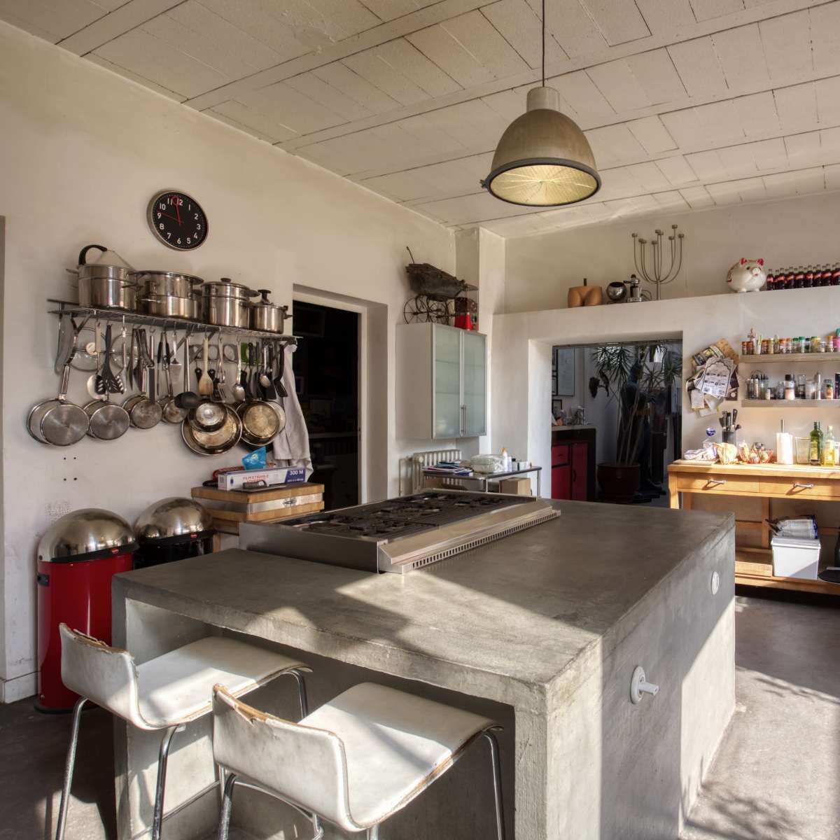 A modern kitchen with a concrete island, stainless steel pots hanging from the wall, and a wooden countertop filled with various cooking ingredients and bottles.