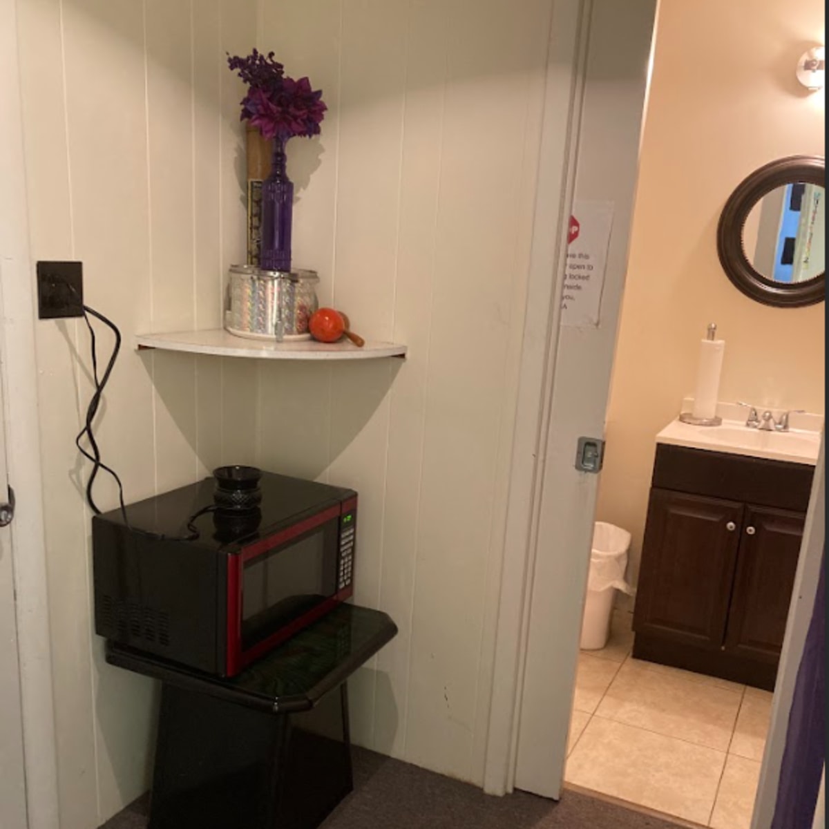 A microwave sits on a small black stand against a white wall, with a shelf above it holding a vase of flowers and an orange, leading to a bathroom visible in the background.
