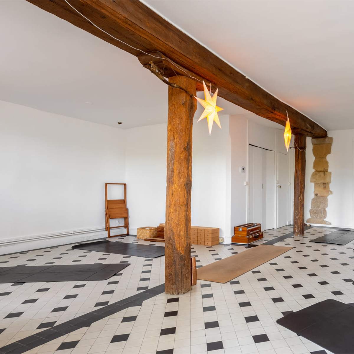 A spacious, minimally furnished room with wooden beams, black and white tile flooring, and several yoga mats arranged for use.
