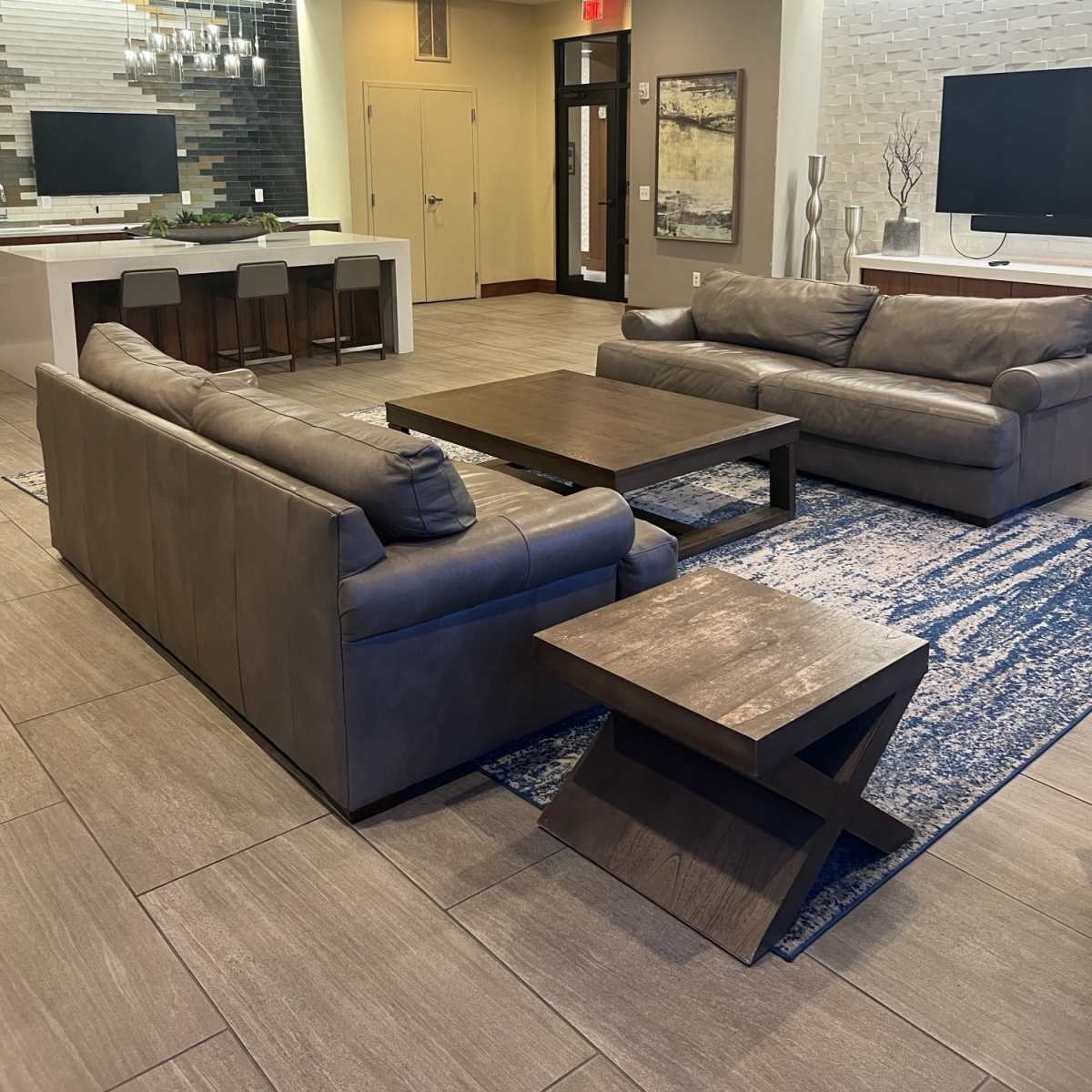 A modern lounge area featuring two gray leather sofas, a wooden coffee table, and a small side table, all set on a blue and white patterned rug.