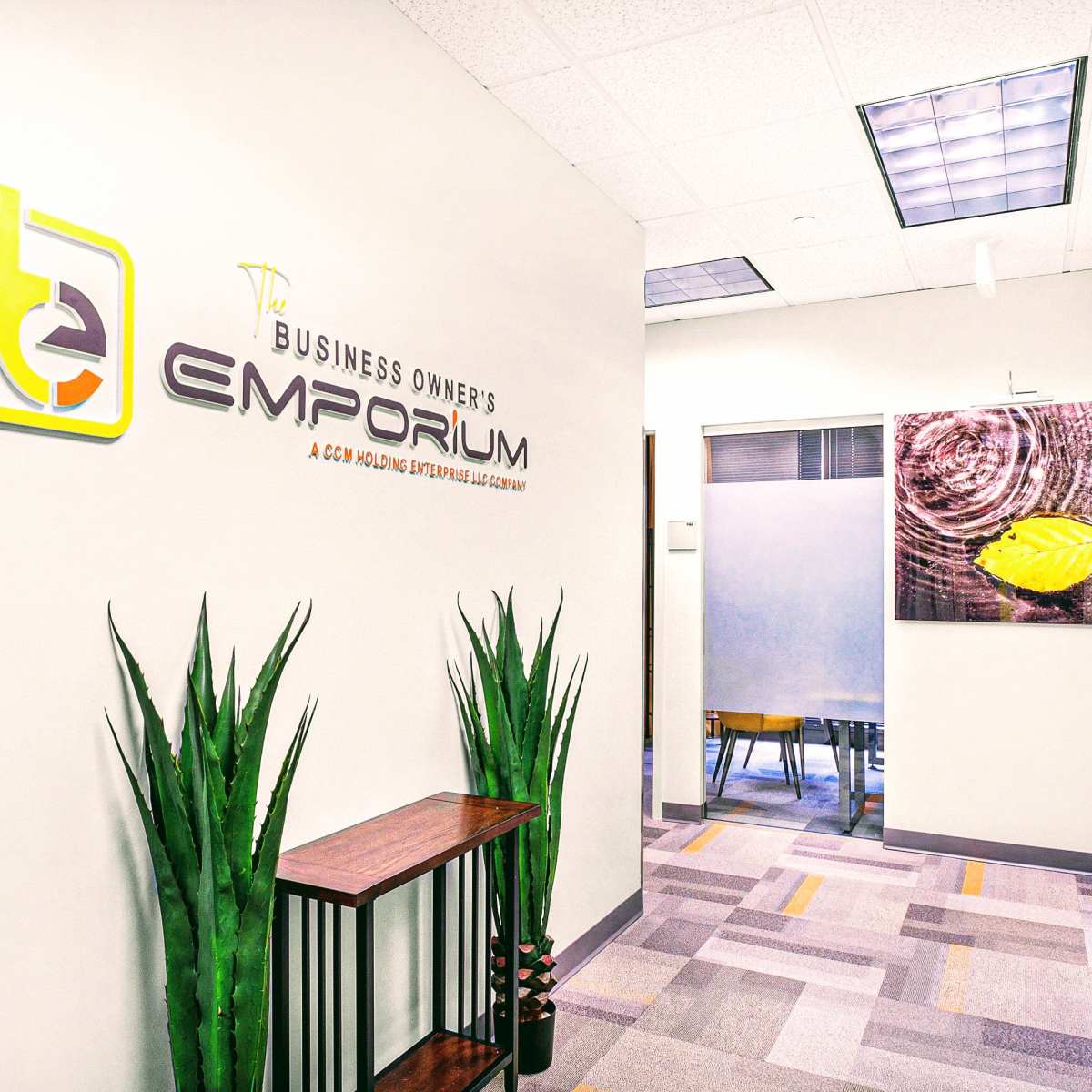 A well-lit office hallway featuring a sign for "The Business Owner's Emporium," along with contemporary decor and plants.