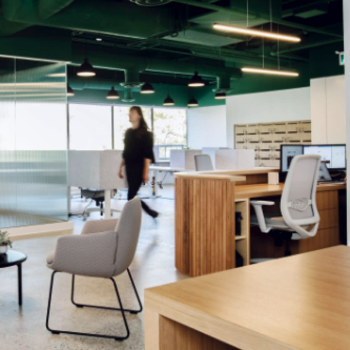 A modern office space with two gray chairs and a small table in the foreground, while a person walks past in the background amidst desks and glass partitions.