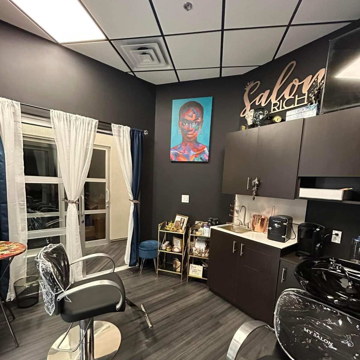 A modern salon interior featuring a styling chair, a washing station, and a colorful artwork on the wall, with curtains framing a window.