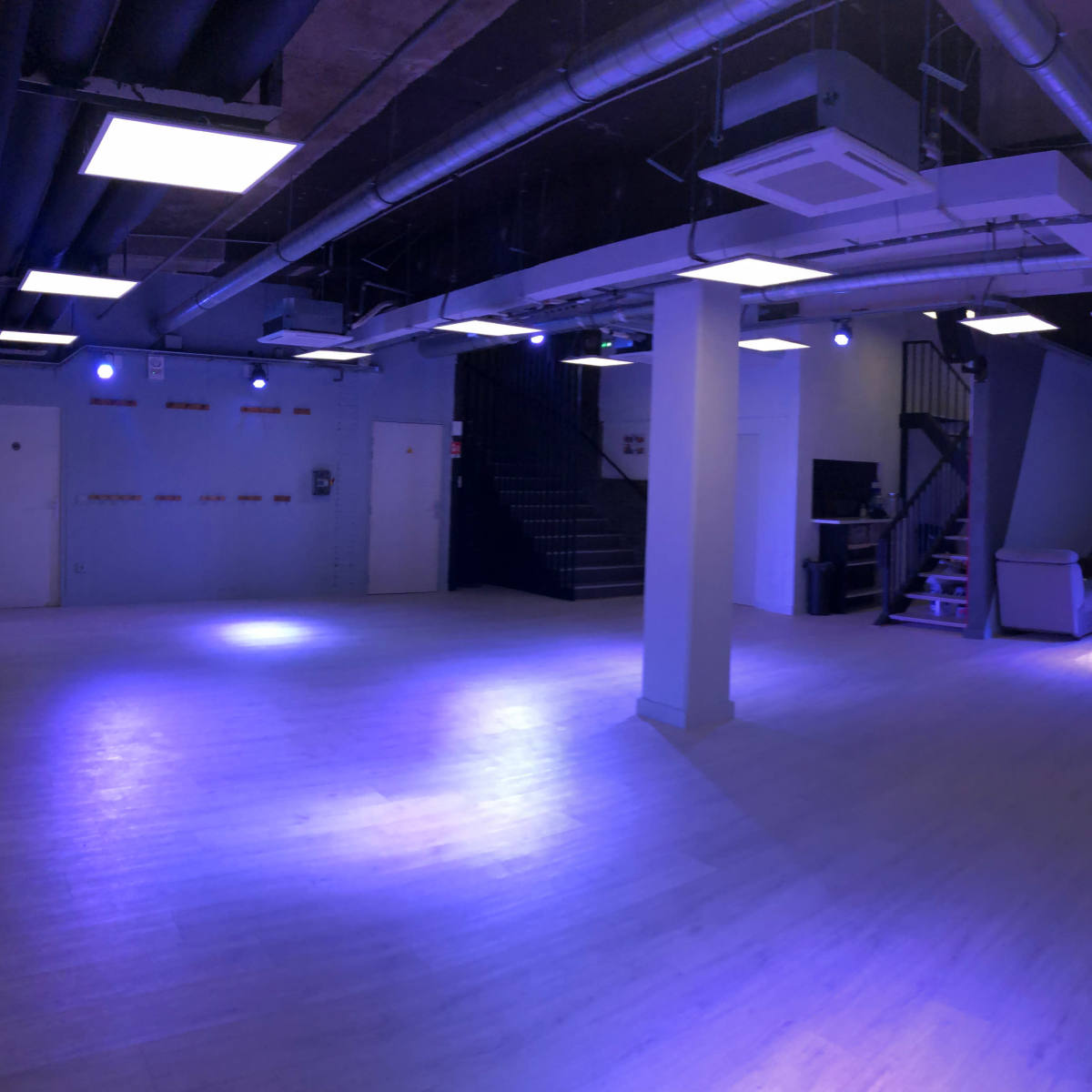 A spacious, dimly lit room with a light blue hue, featuring a staircase at one end and various seating arrangements along the walls.