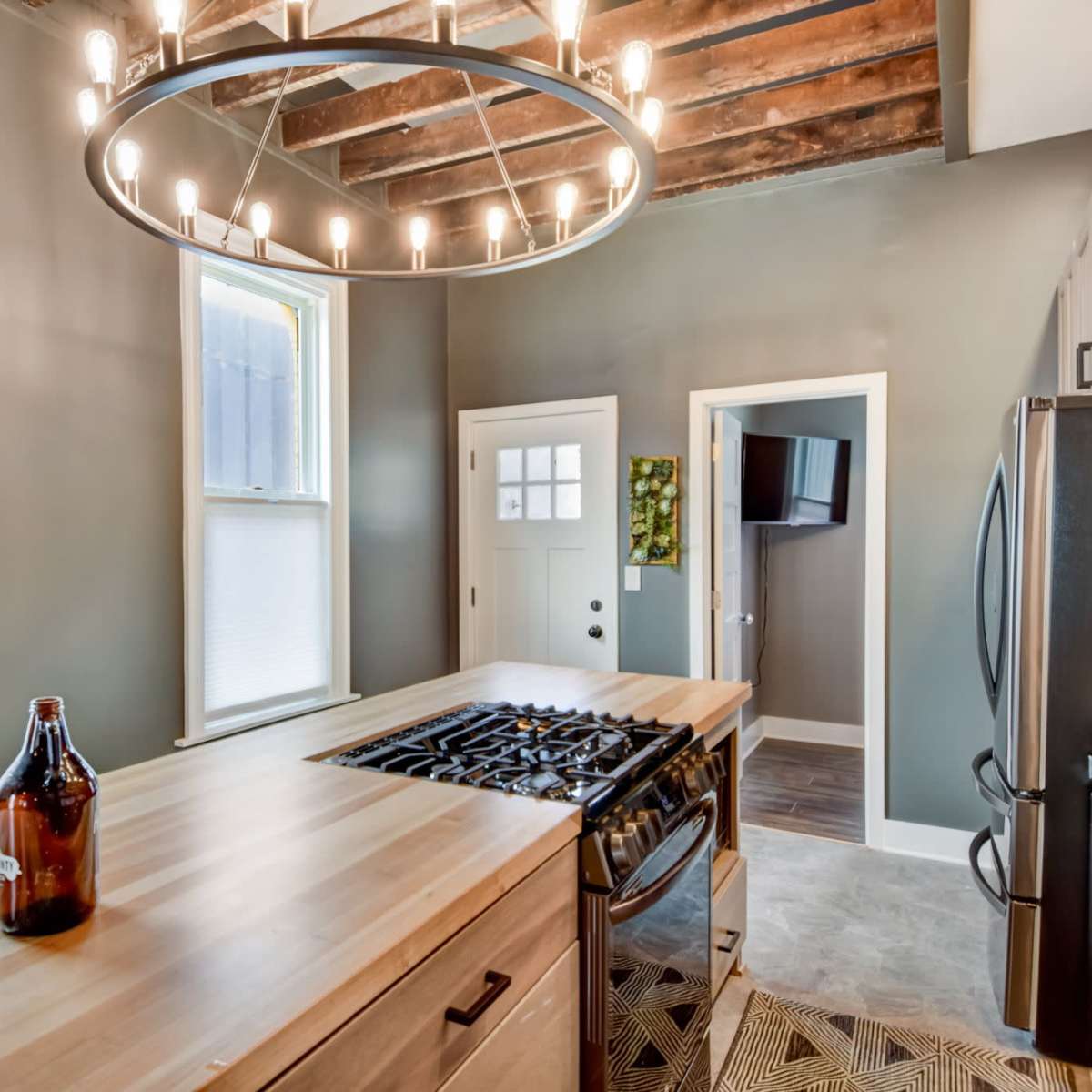 A modern kitchen features a central wooden island with a gas stove and a large circular chandelier above, complemented by exposed wooden beams and gray walls.