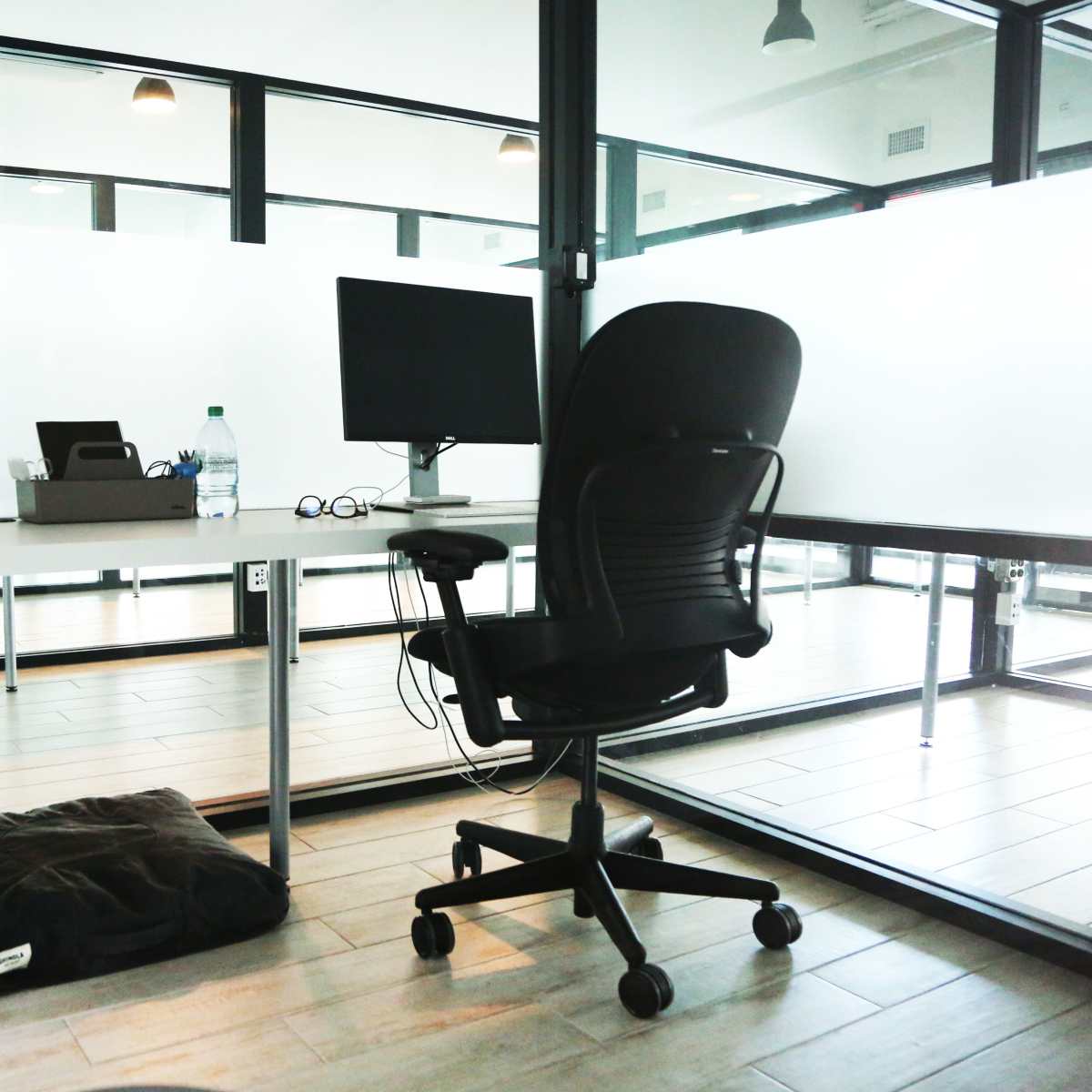 An empty office space features a desk with a computer monitor, a chair, and a small plant, surrounded by glass walls.