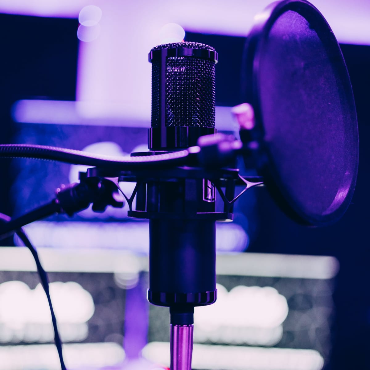 A microphone with a pop filter is positioned in front of computer screens illuminated by purple lighting.