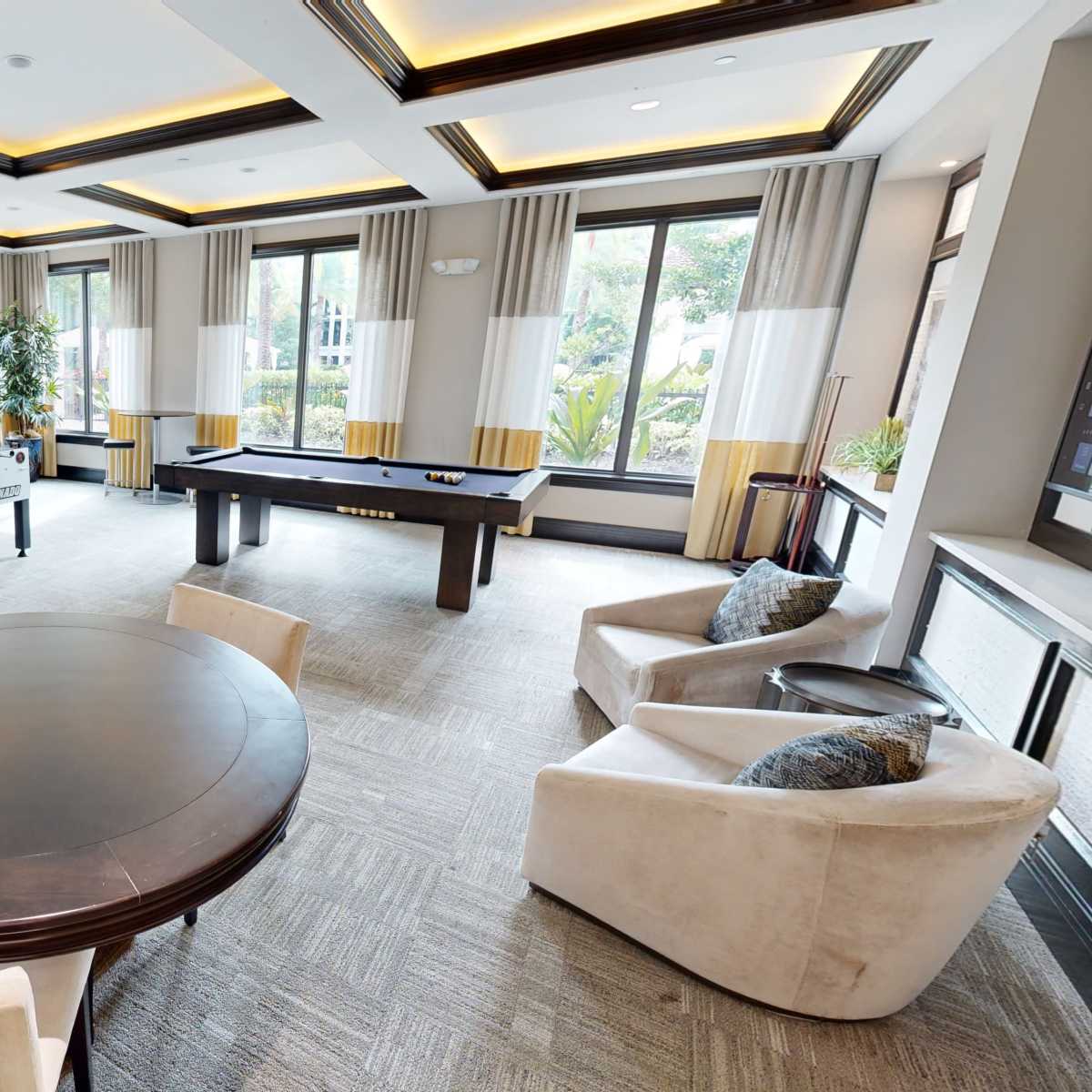 A modern lounge area with a round table, plush chairs, a foosball table, and large windows allowing natural light.