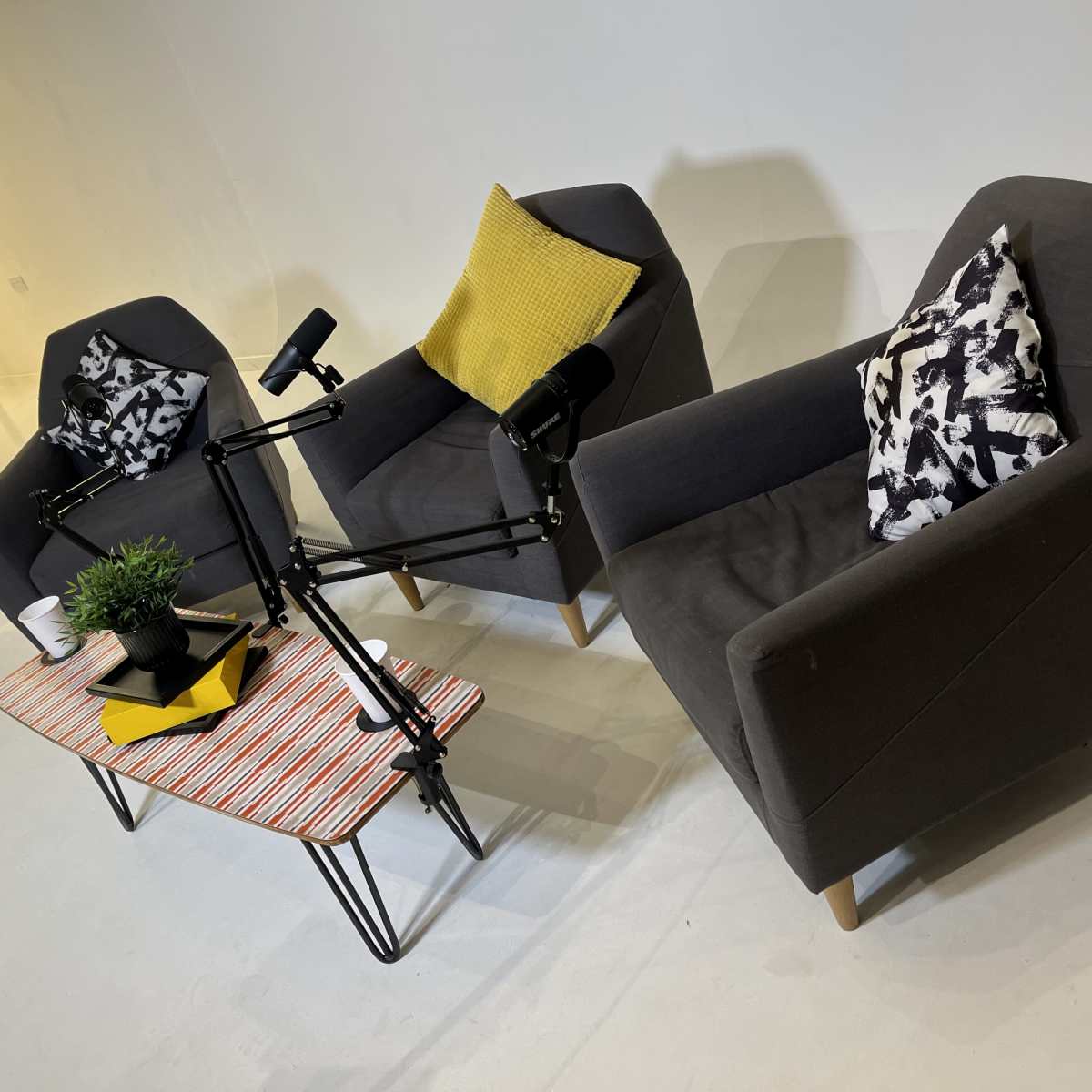 Two gray armchairs with patterned cushions positioned around a small, colorful coffee table featuring a plant and two microphone stands.